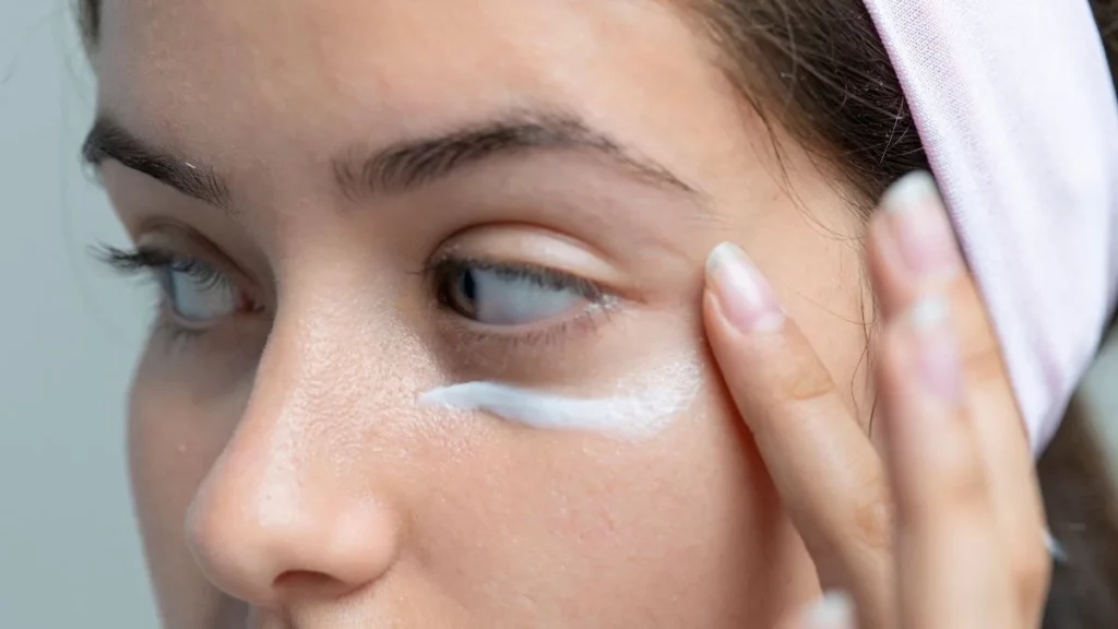 mulher aplicando creme na área dos olhos com dedos enquanto cuida da pele do rosto focando em hidratação e redução de olheiras