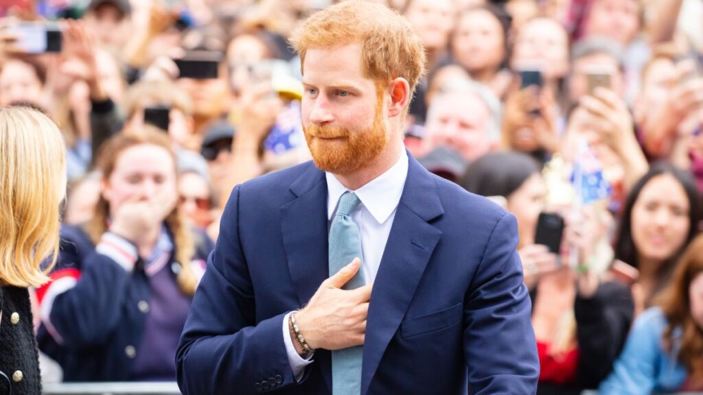 Príncipe Harry de terno azul cumprimenta o público durante evento com multidão ao fundo