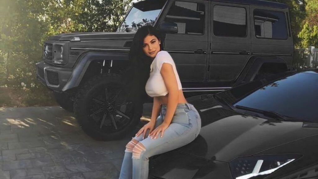 Vida luxuosa das celebridades: como vivem os famosos mais ricos Kylie Jenner posando sentada em carro esportivo de luxo com SUV ao fundo em cenário externo durante o dia, mostrando a vida luxuosa das celebridades