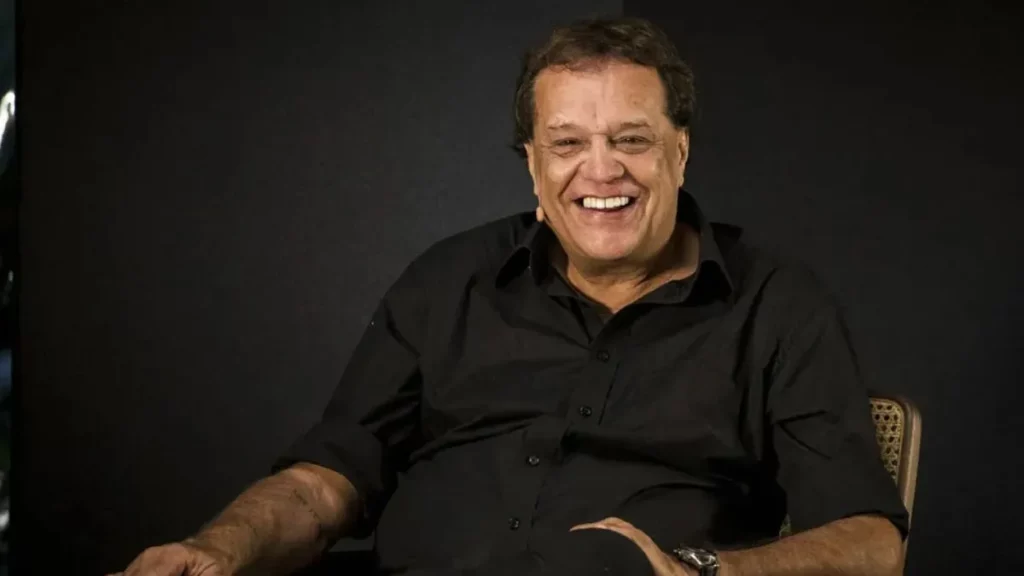 Atores da Globo que morreram recentemente: quem eram, trajetórias e principais obras 3 Dennis Carvalho, homem sorridente com camisa preta, posa sentado diante de fundo escuro em retrato iluminado.