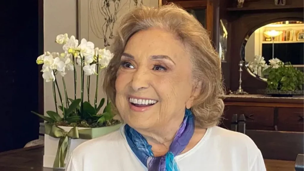 Atores da Globo que morreram recentemente: quem eram, trajetórias e principais obras 9 Eva Wilma sorrindo em ambiente interno, com lenço azul no pescoço e flores brancas ao fundo.