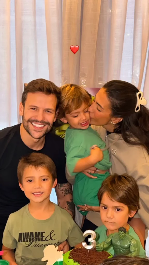 Gabi Brandt e Saulo Poncio aparecem sorrindo ao lado dos três filhos em celebração familiar dentro de casa.