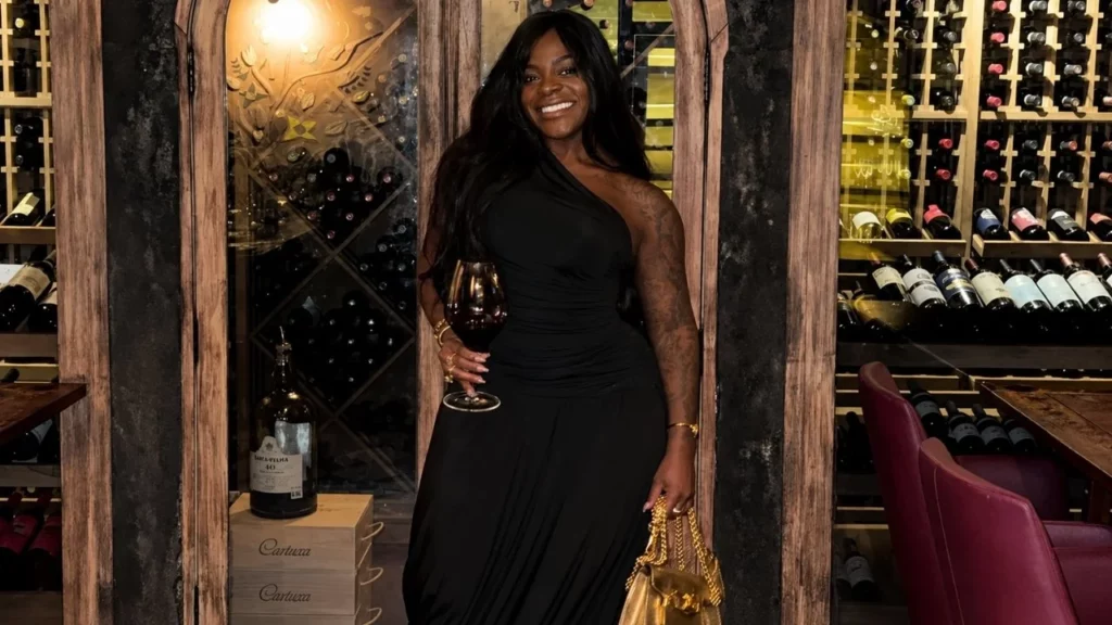 Jojo Todynho posa sorrindo com vestido preto e taça em adega.