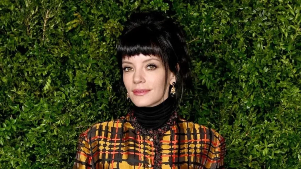 Lily Allen, mulher com cabelo preto preso e franja, usa vestido xadrez diante de parede coberta por folhagens verdes.