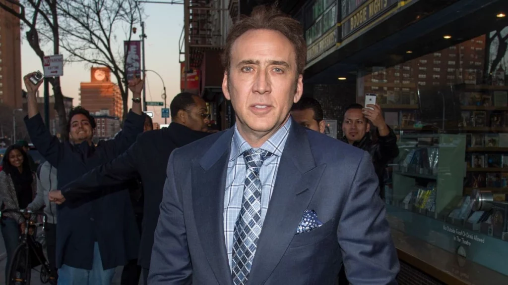 Nicolas Cage posa de terno em rua movimentada enquanto fãs aparecem ao fundo tirando fotos e acenando.