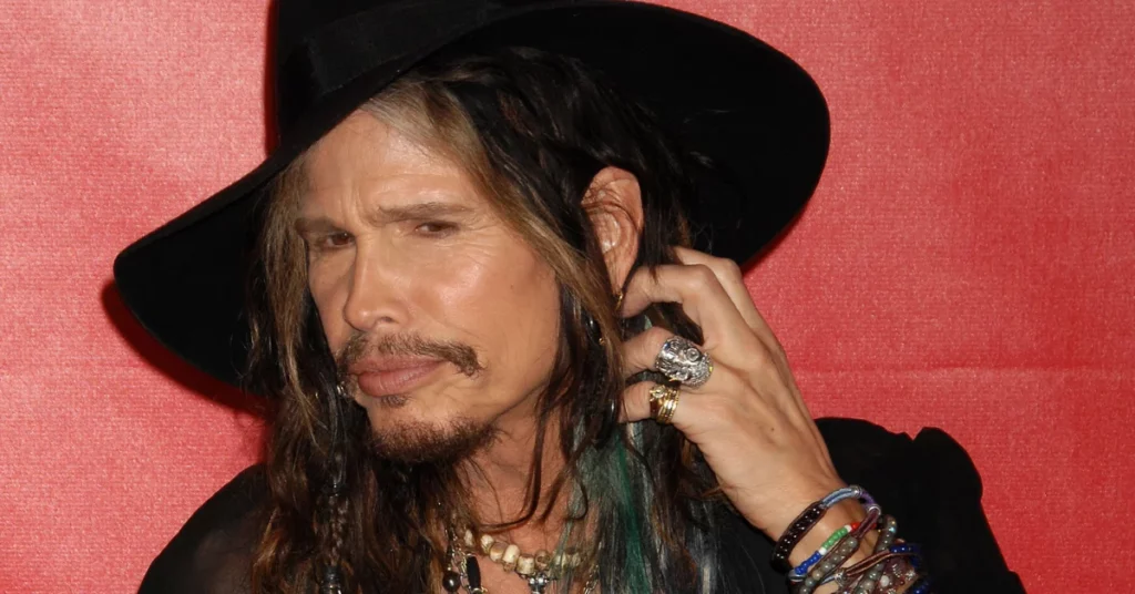 Steven Tyler aparece de chapéu preto, joias e expressão séria diante de fundo vermelho em foto usada em matéria sobre processo judicial