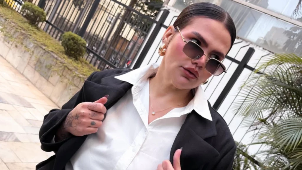 Thais Carla revela próximo passo após perder 101 kg e faz promessa: ‘Não vão reconhecer’ Thais Carla de camisa branca, blazer preto e óculos escuros posa ao ar livre em área urbana com vegetação e grades ao fundo.