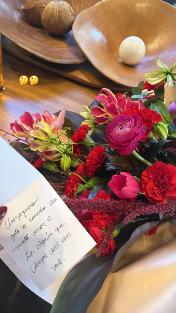 Buquê de flores vermelhas e rosas aparece ao lado de cartão escrito à mão enviado anonimamente para Ana Castela