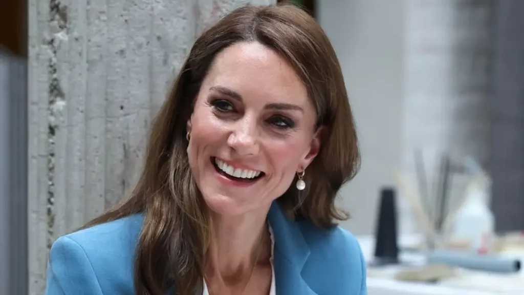 Kate Middleton sorri durante evento oficial, usando blazer azul-claro e brincos de pérola.