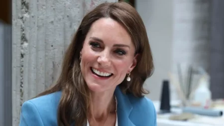 Kate Middleton sorri durante evento oficial, usando blazer azul-claro e brincos de pérola.