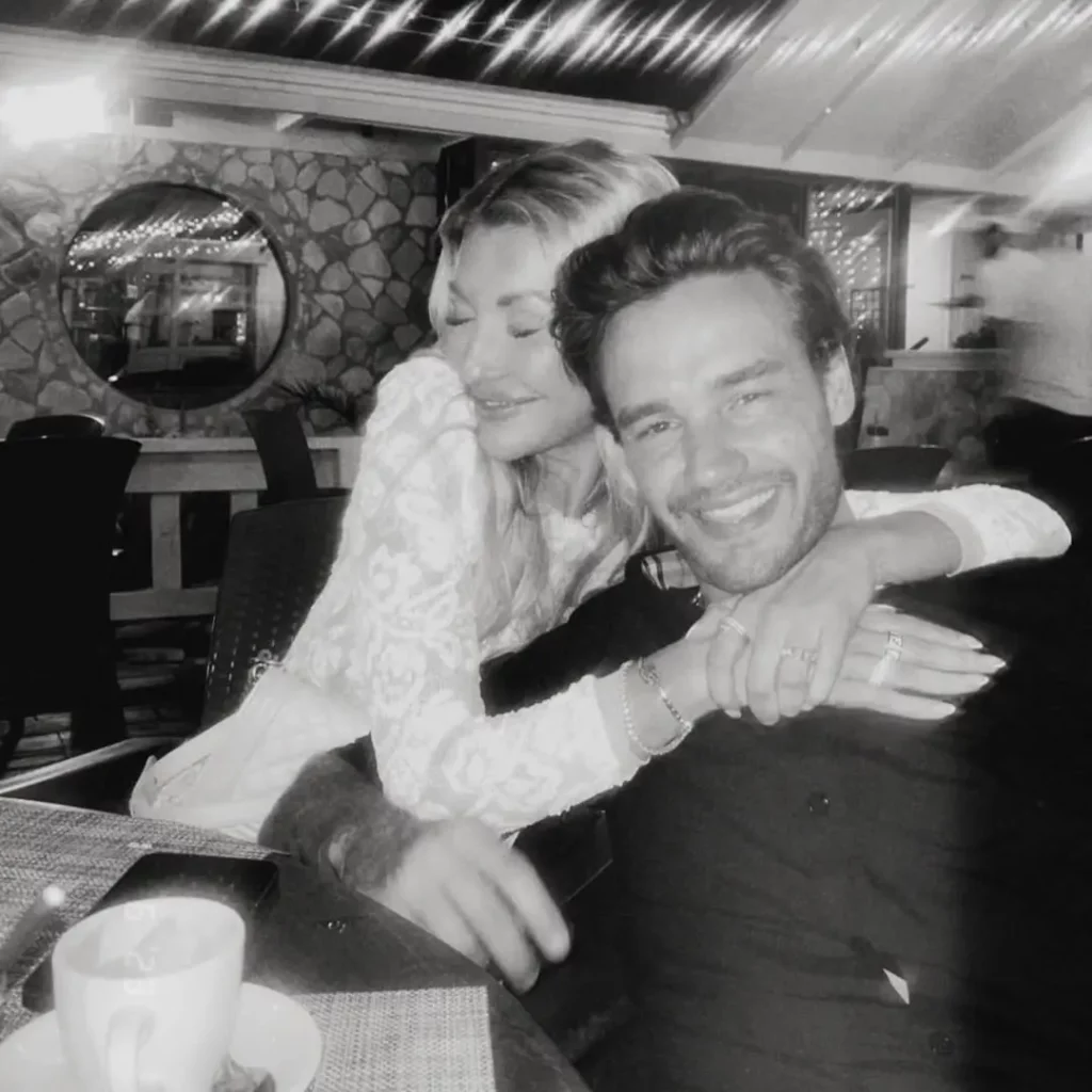 Foto em preto e branco mostra Kate Cassidy abraçando Liam Payne durante um jantar em ambiente iluminado.