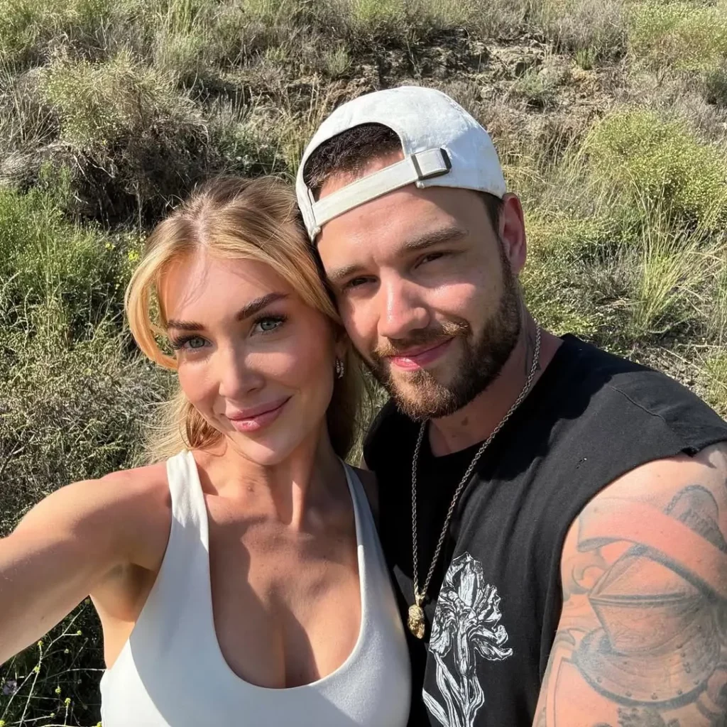 Kate Cassidy e Liam Payne tiram uma selfie juntos em uma área de natureza com vegetação ao fundo.