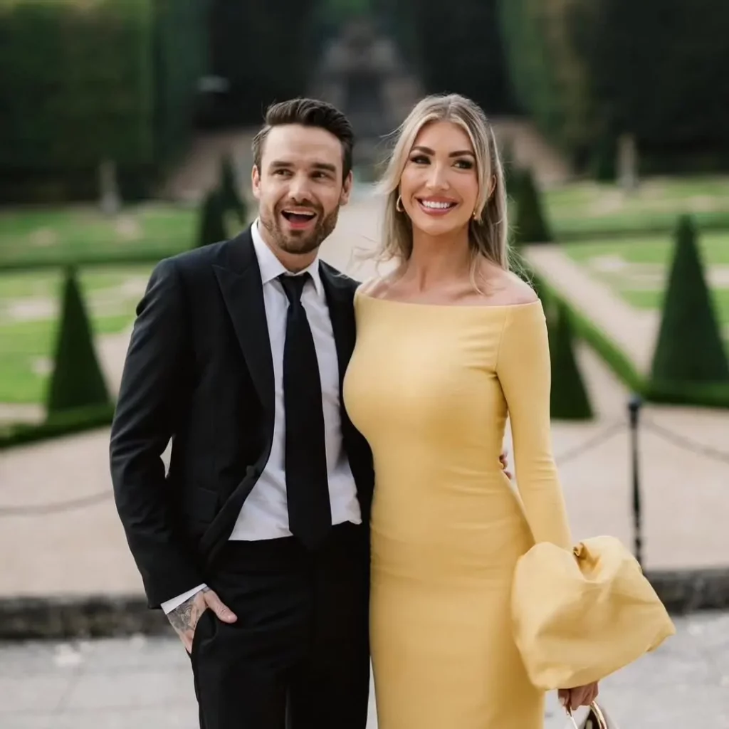 Liam Payne e Kate Cassidy posam sorrindo durante um evento ao ar livre, usando trajes elegantes.