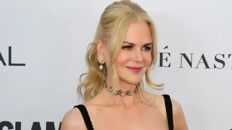 Nicole Kidman posa sorrindo em evento de tapete vermelho usando vestido preto e acessórios prateados.