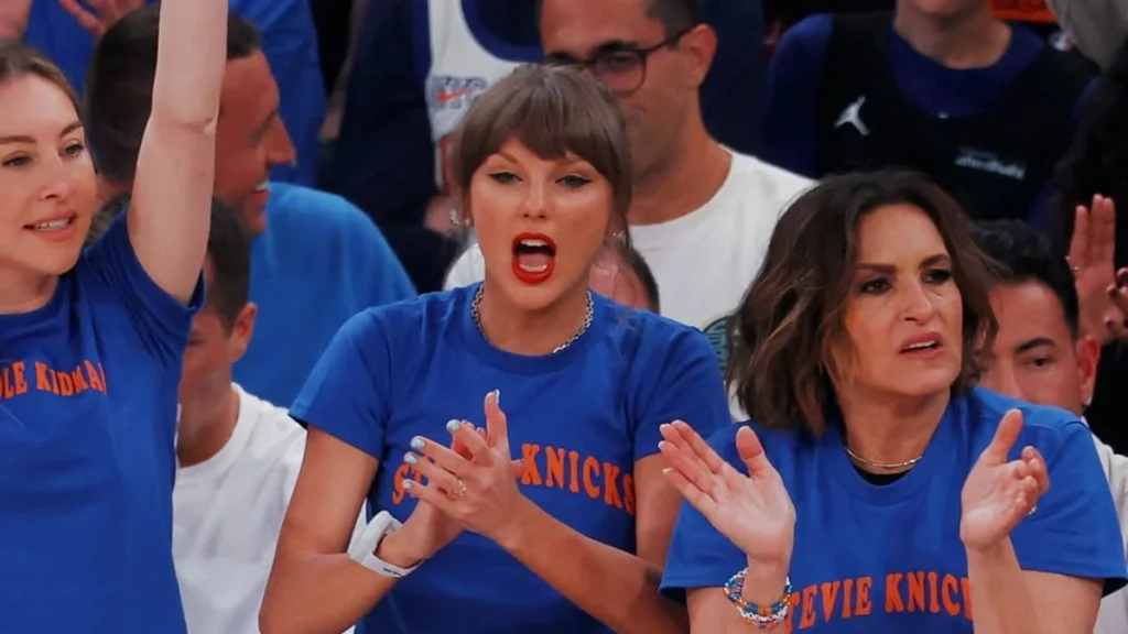 Taylor Swift comemora e reage com entusiasmo durante partida de basquete, usando camiseta azul nas arquibancadas e interagindo com amigos e torcedores.
