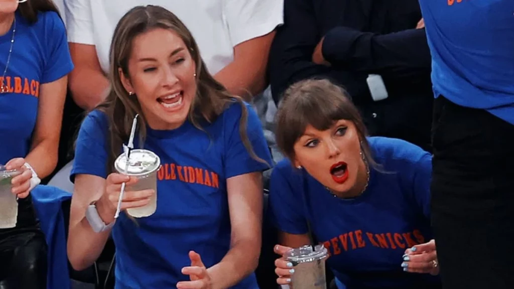 Taylor Swift comemora e reage com entusiasmo durante partida de basquete, usando camiseta azul nas arquibancadas e interagindo com amigos e torcedores.
