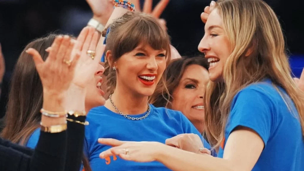 Taylor Swift comemora e reage com entusiasmo durante partida de basquete, usando camiseta azul nas arquibancadas e interagindo com amigos e torcedores.