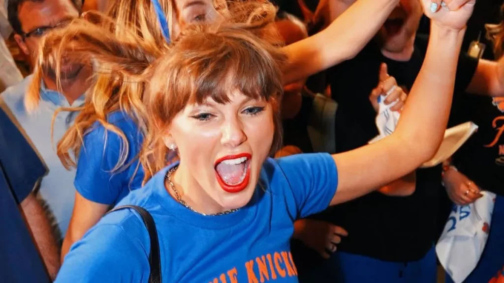 Taylor Swift comemora e reage com entusiasmo durante partida de basquete, usando camiseta azul nas arquibancadas e interagindo com amigos e torcedores.