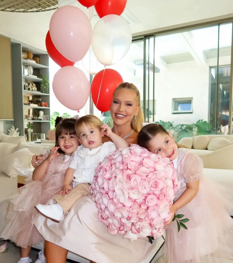 Virginia Fonseca posa sorrindo com os três filhos dentro de casa, segurando um grande buquê de flores rosas enquanto balões decorativos aparecem ao fundo.