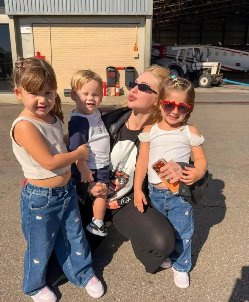 Virginia Fonseca posa ao ar livre com Maria Alice, Maria Flor e José Leonardo em uma área próxima a um hangar de aeronaves.