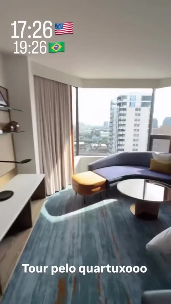 Quarto da suíte conta com sofá curvo, poltronas, mesa de centro e grandes janelas com vista urbana.