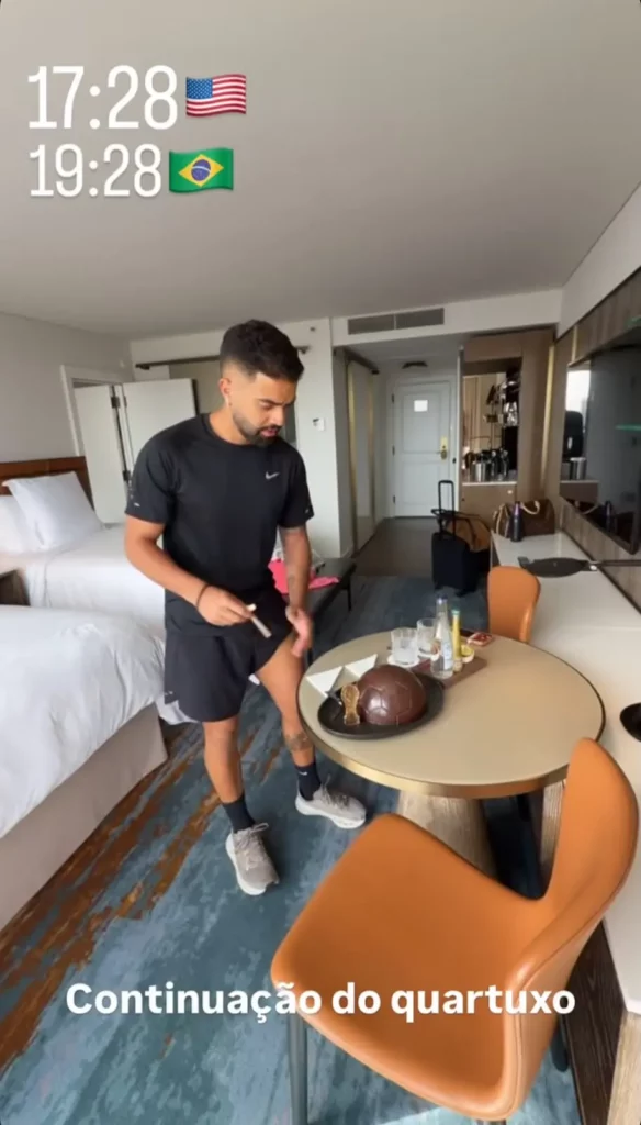 Homem observa um mimo em formato de bola de futebol sobre a mesa do quarto, que possui duas camas de casal e decoração contemporânea.