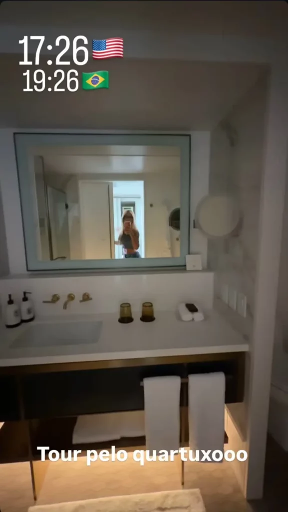 Virginia faz selfie diante do espelho enquanto exibe o banheiro da suíte do Four Seasons Hotel Houston, com bancada de mármore, pia e acabamentos em dourado.