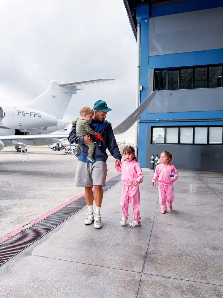 Zé Felipe caminha em um hangar segurando José Leonardo no colo enquanto acompanha Maria Alice e Maria Flor.