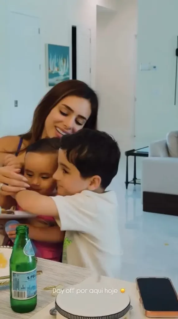 Bruna Biancardi com a filha em um ambiente interno. Na mesa à frente, há uma garrafa verde de água e um celular. Texto sobreposto: "Day off por aqui hoje ☀️".