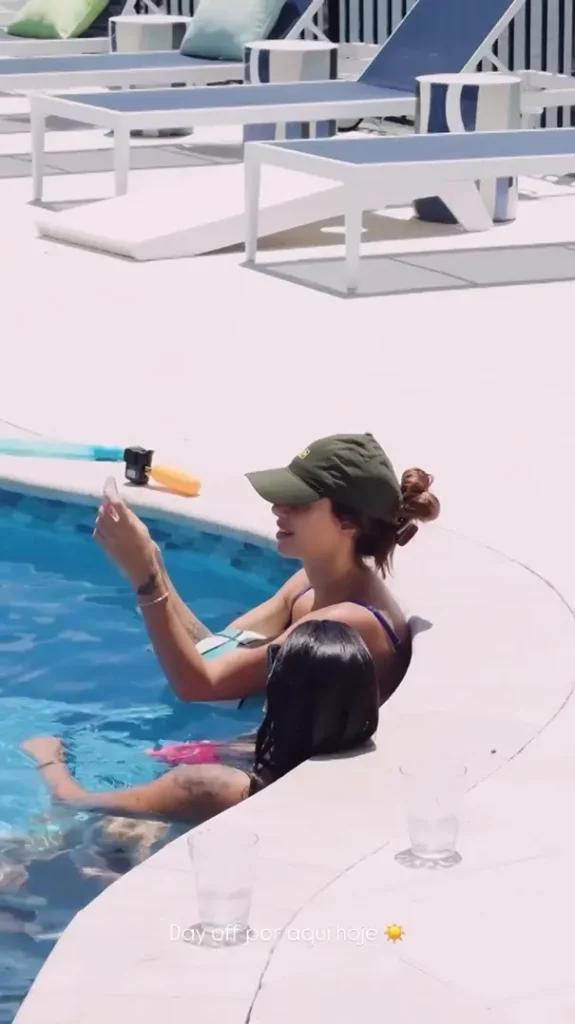 Bruna Biancardi relaxando na beira de uma piscina em um dia ensolarado; uma delas usa boné verde e segura um celular. Ao fundo, espreguiçadeiras. Texto sobreposto: "Day off por aqui hoje ☀️".