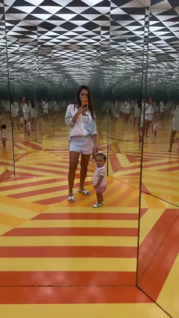 Bruna Biancardi e filha em pé em uma sala de espelhos com um padrão de listras laranja e amarelo no chão.