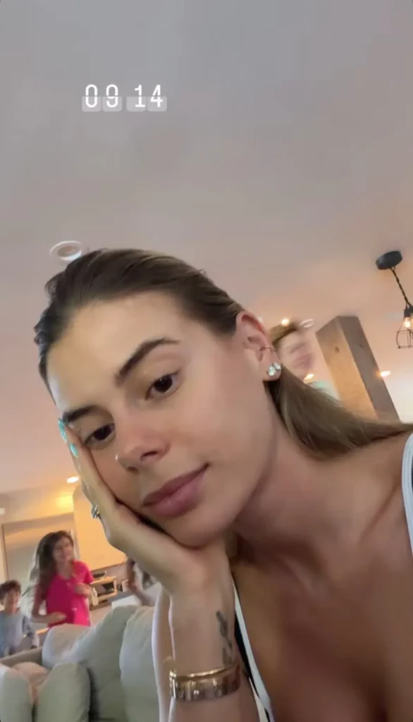 Carol Cabrino posa para uma selfie, apoiando o rosto na mão. Uma criança em um top rosa é visível ao fundo, fora de foco.