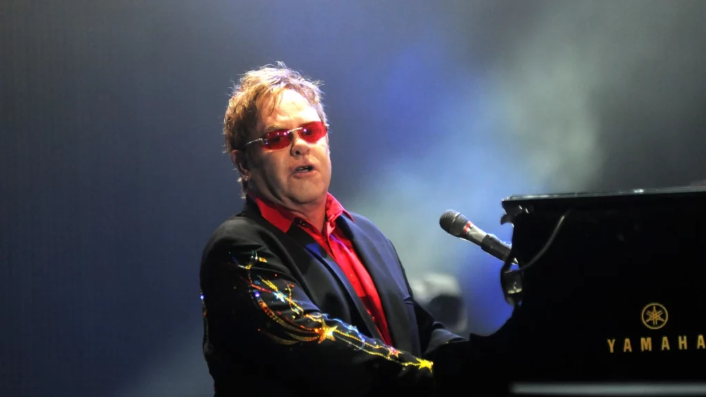 Elton John se apresenta no palco usando óculos vermelhos e blazer escuro, sob iluminação de show.