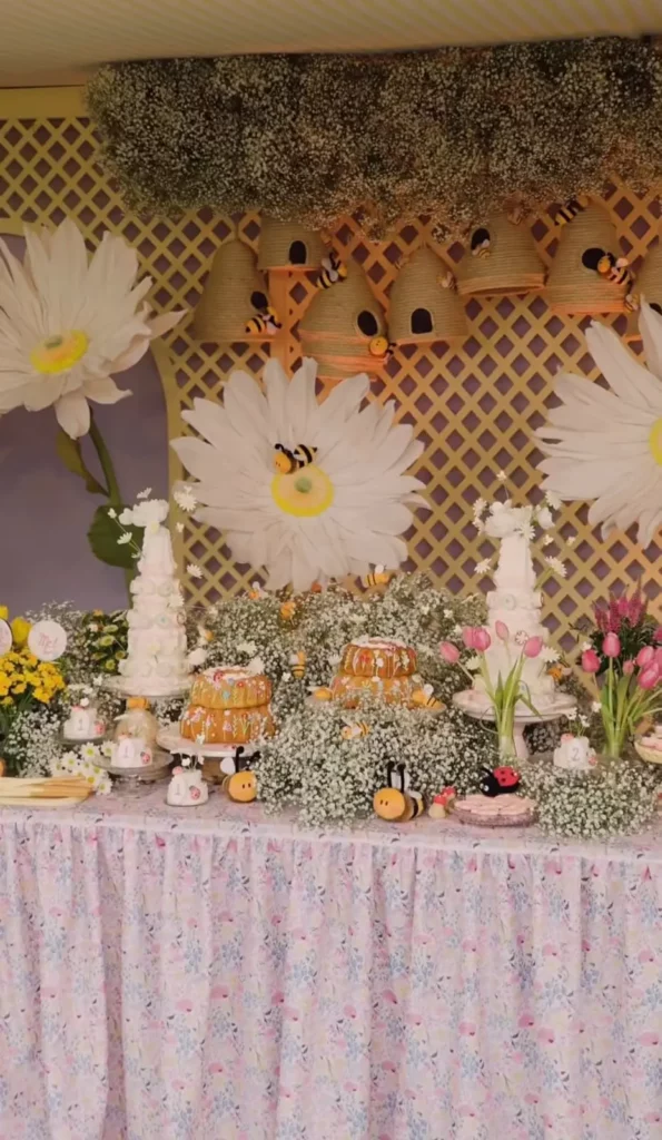 ma visão ampla de toda a mesa de sobremesas principal. O fundo é a treliça de madeira com margaridas gigantes de papel e colmeias de corda penduradas com abelhas de pelúcia. A mesa tem uma toalha floral e está coberta por arranjos de gipsofila e outras flores. Há dois bolos principais (Helena à direita, Mel à esquerda) e arranjos de tulípas rosa entre eles. Pelúcias de abelha e joaninha estão espalhadas.