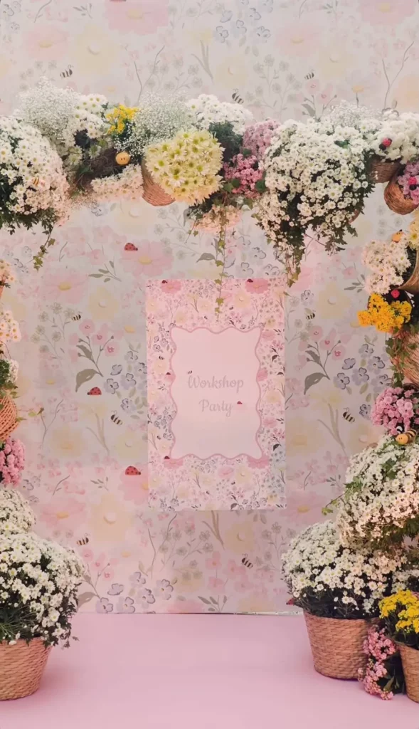 O cenário da comemoração, com um arco decorativo feito de muitas gipsofilas brancas e outras flores coloridas. No centro, um banner rosa pastel pendurado com um padrão floral e o texto "Workshop Party" em fonte cursiva. O fundo é um papel de parede floral pastel com cestas de vime na base.