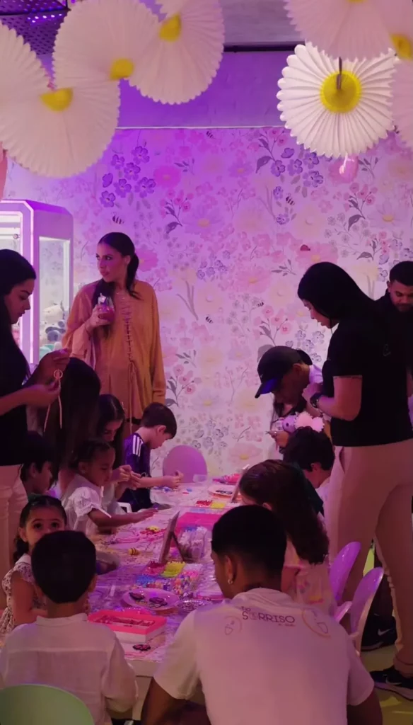 Cena mais ampla da comemoração, onde várias crianças e adultos estão sentados em mesas de atividades, criando artesanatos coloridos. A decoração ao fundo é o mesmo papel de parede floral/jardim, com grandes margaridas penduradas e iluminação rosa suave.