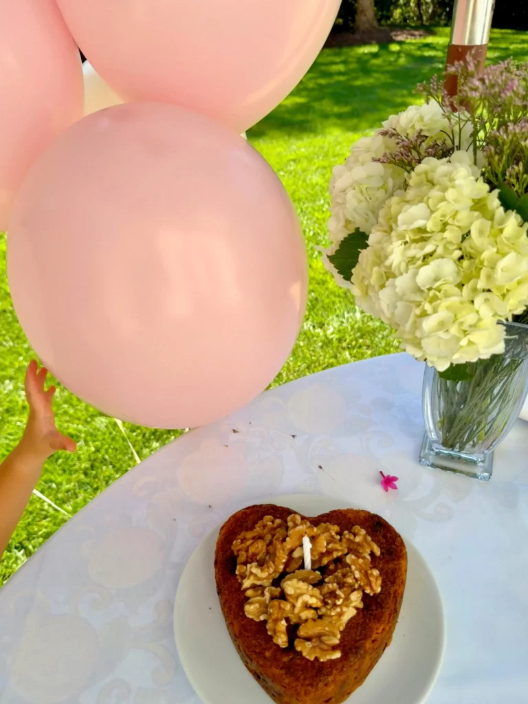 Mesa da comemoração de aniversário de Gisele Bündchen com bolo em formato de coração decorado com nozes, flores brancas e balões em tons pastel.