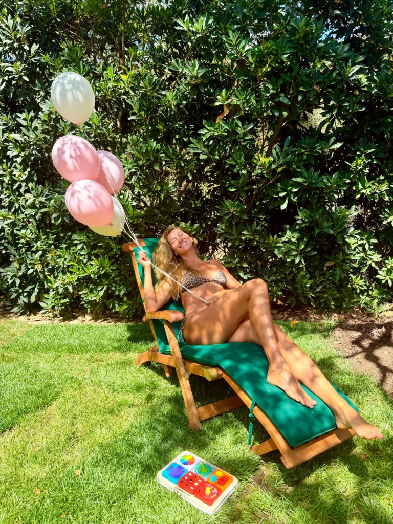 Gisele Bündchen posa sorrindo em uma espreguiçadeira no jardim, usando biquíni de animal print e segurando balões em tons de rosa e branco.