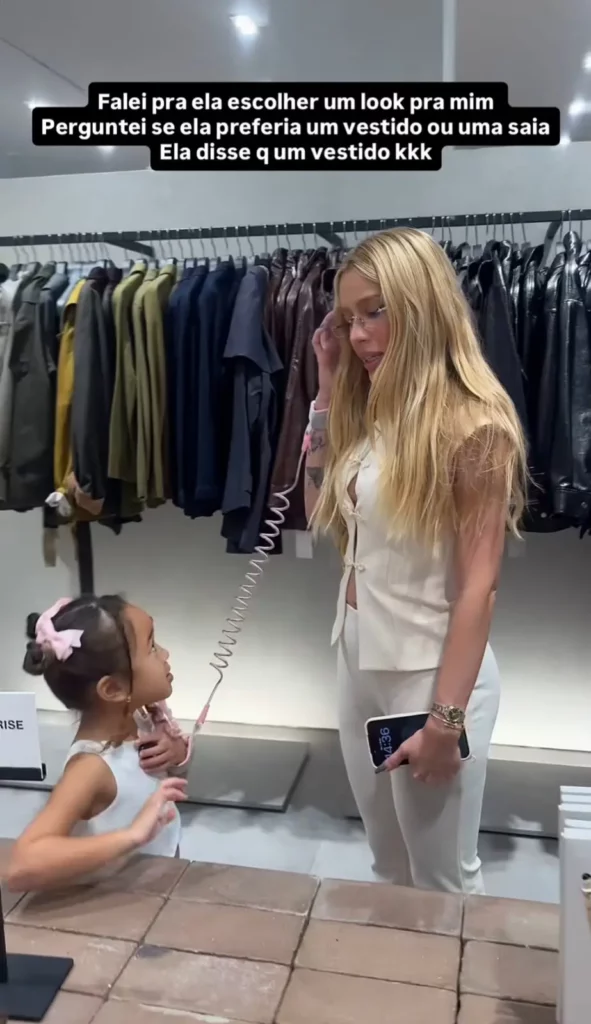 Karoline Lima e sua filha se olhando em uma loja de roupas. Há texto em português sobreposto no topo da imagem.