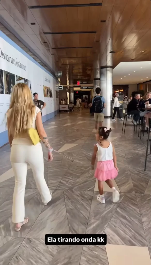 Karoline Lima, vista de costas vestindo conjunto branco e bolsa amarela, caminha por um corredor de shopping ao lado de sua filha, Cecília, que usa uma saia rosa. As duas estão conectadas por uma pulseira guia de segurança infantil. Texto sobreposto na parte inferior: "Ela tirando onda kk".