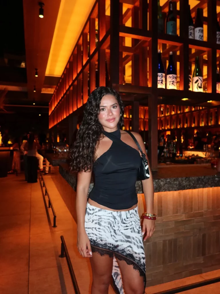 Maisa posa em um restaurante durante a noite, usando blusa preta e saia estampada, com prateleiras iluminadas ao fundo.
