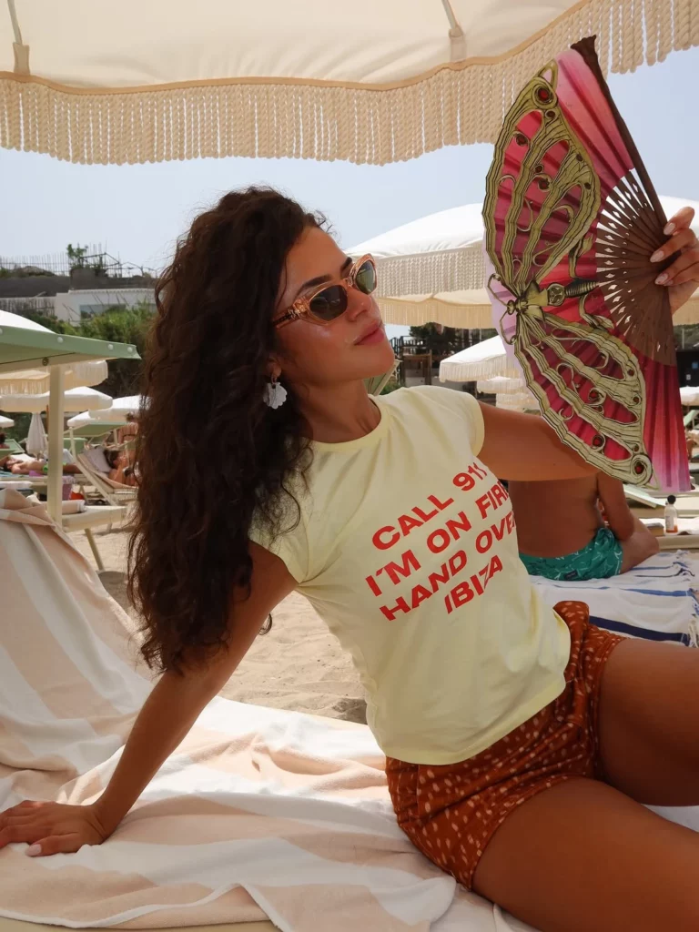 Maisa segura um leque colorido enquanto relaxa em uma espreguiçadeira de praia, usando camiseta amarela e óculos de sol.
