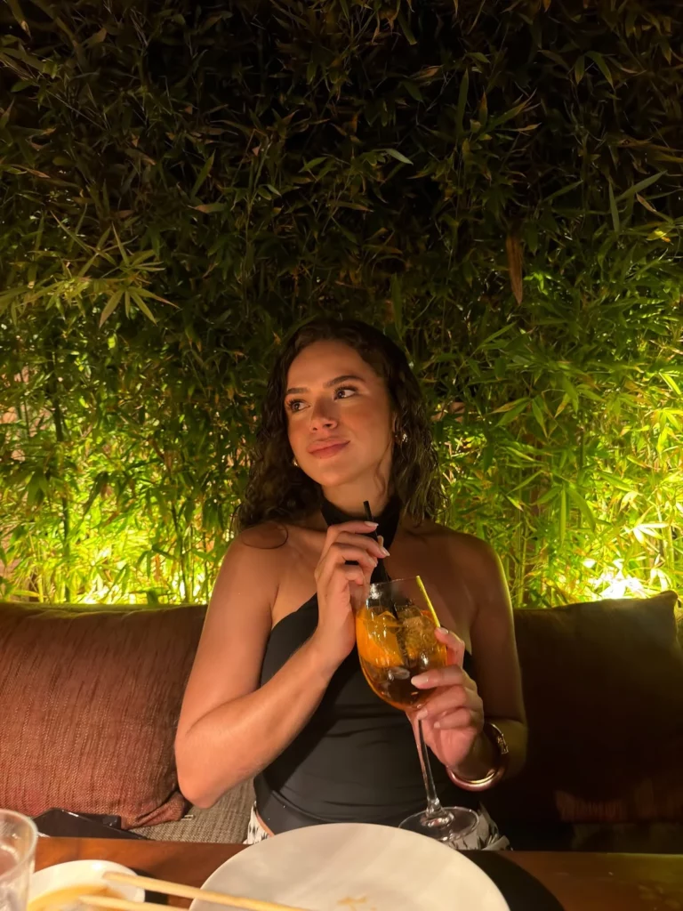 Maisa segura uma taça de bebida enquanto está sentada em um restaurante ao ar livre cercado por plantas e iluminação aconchegante.