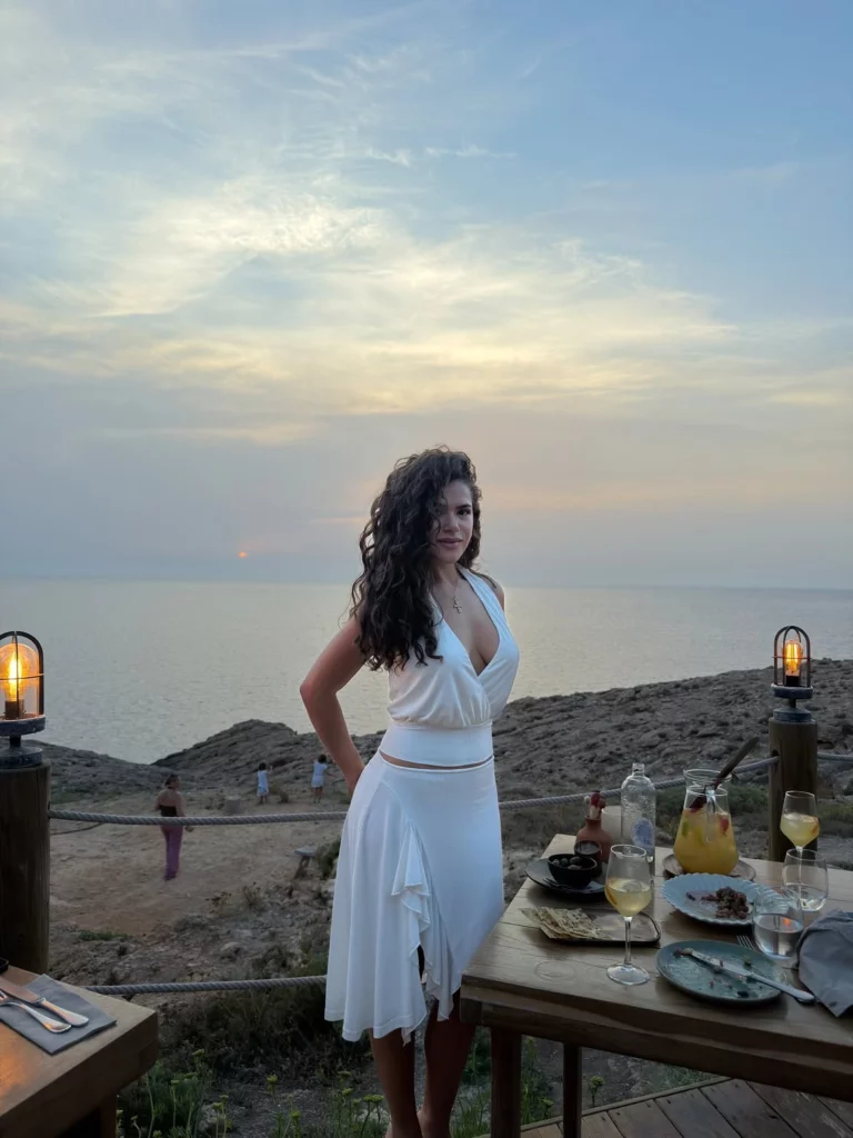 Maisa posa usando um vestido branco durante um jantar ao pôr do sol, com vista para o mar e uma mesa posta ao lado.
