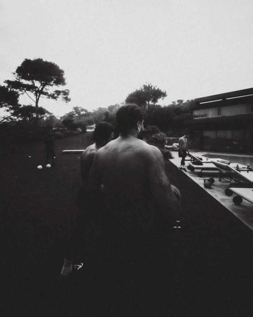 Memphis Depay aparece de costas ao lado de outra pessoa em uma área externa de uma mansão, com piscina e espreguiçadeiras ao fundo, em foto em preto e branco.