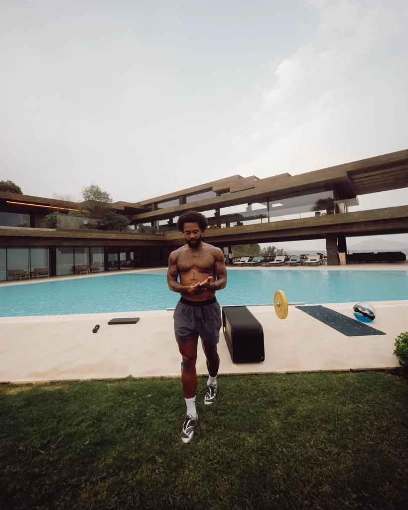 Memphis Depay caminha sem camisa em frente à piscina de uma mansão, com equipamentos de treino espalhados pelo gramado.
