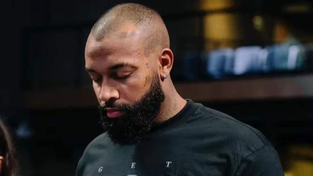 Nego Di, de cabeça raspada e barba cheia, olhando para baixo, usando camiseta preta em ambiente interno desfocado.