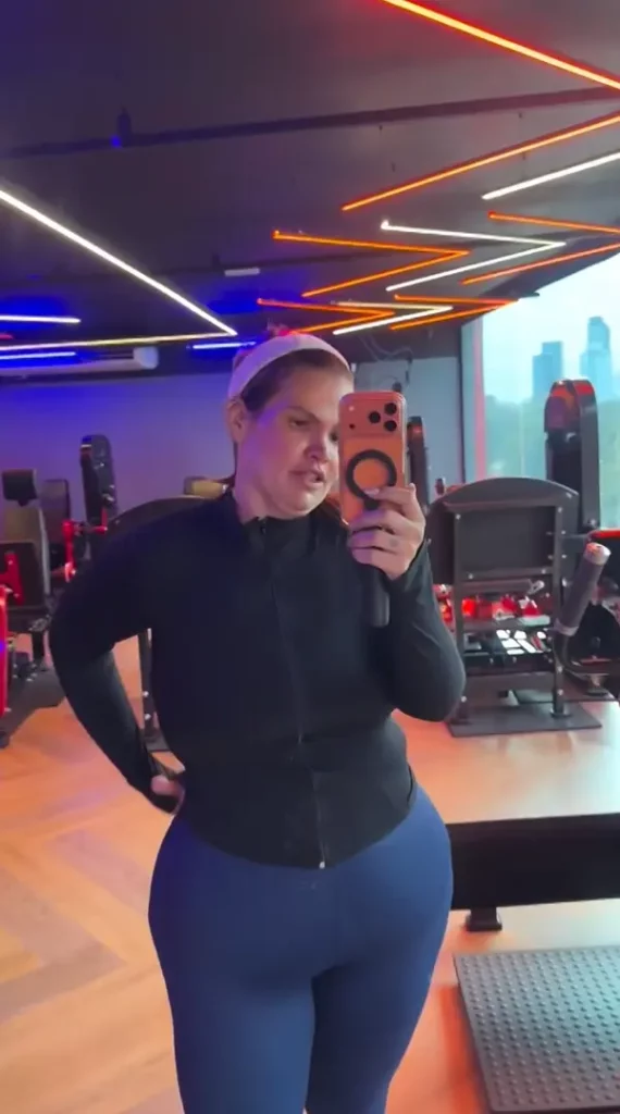 Após perder mais de 100 kg, Thais Carla revela que roupas de academia não servem mais 3 Imagem de corpo inteiro da influenciadora Thais Carla, vestida para treino com jaqueta preta e calças azuis, posando com um telefone na mão em uma academia.