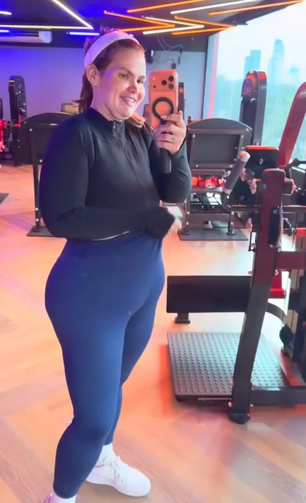 Após perder mais de 100 kg, Thais Carla revela que roupas de academia não servem mais 1 Pose de três quartos de Thais Carla sorrindo com um telefone, em uma academia com vista para a cidade e luzes de neon.
