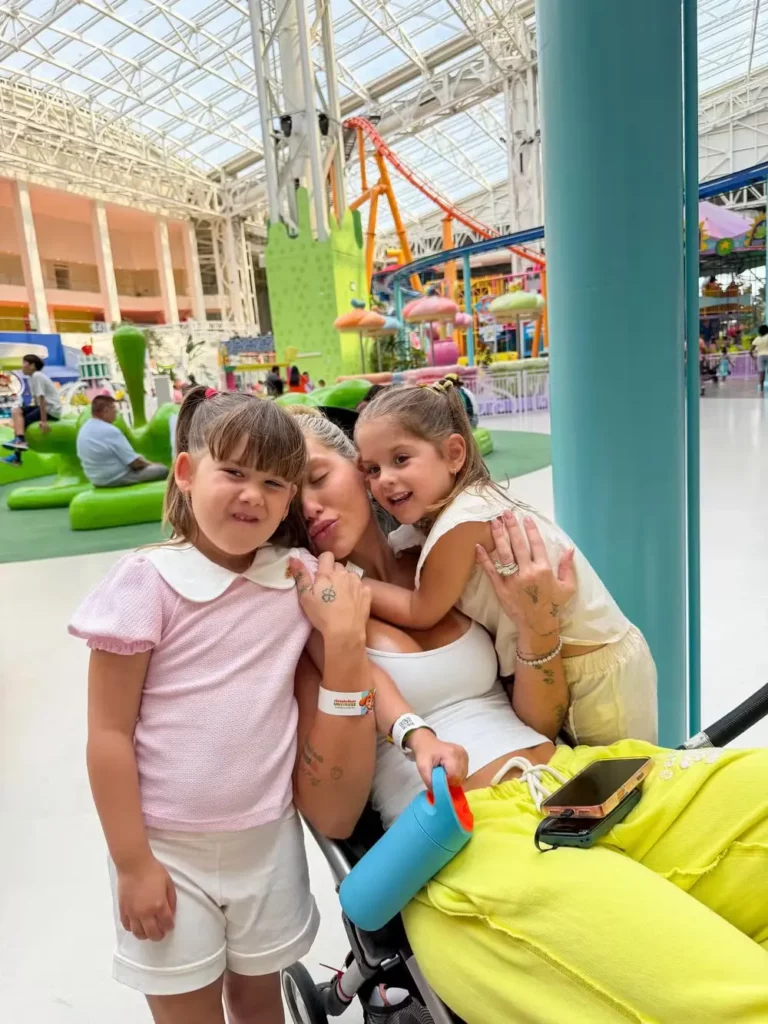 Virginia em um carrinho de bebê, segurando Maria Flor e Maria Alice no parque de diversões coberto Nickelodeon Universe, com uma montanha-russa ao fundo.